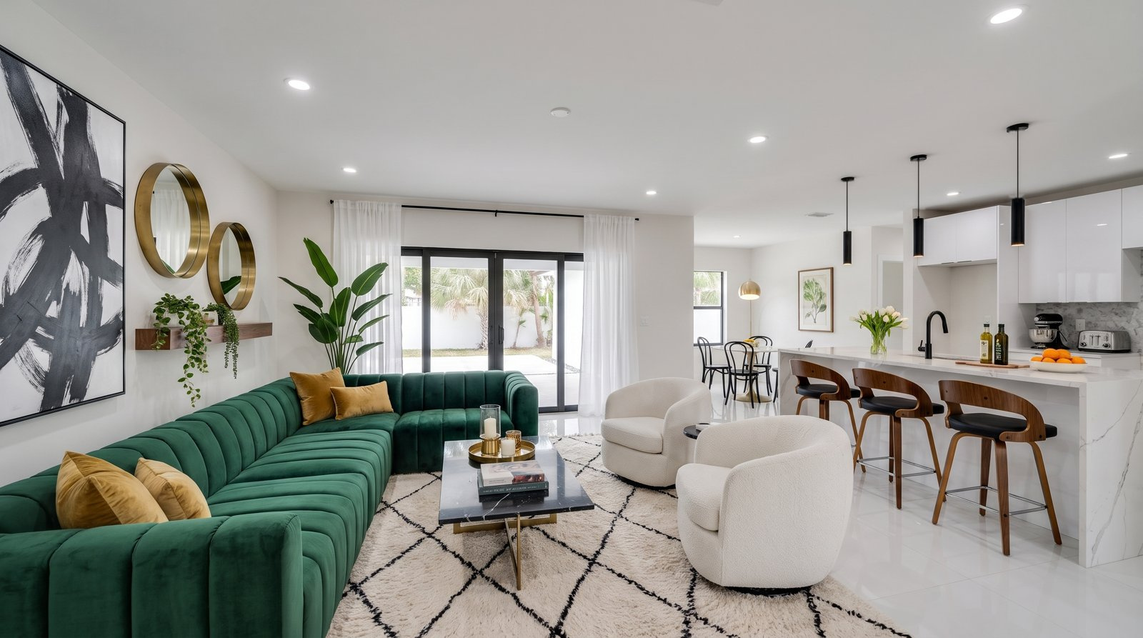 Open living room and kitchen staged with an emerald velvet sectional, marble coffee table and brass mirrors
